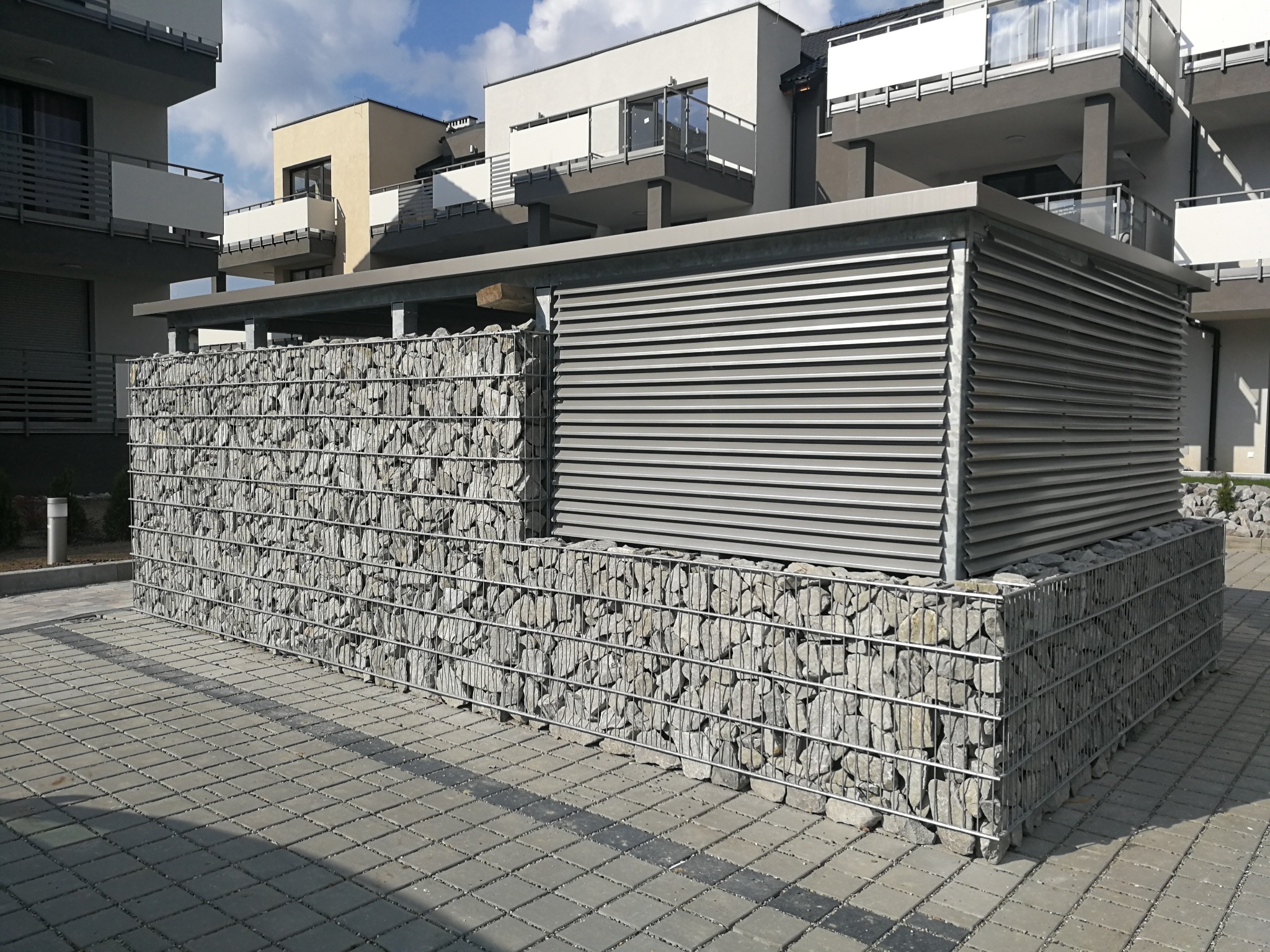 Gabionowa konstrukcja z szarego kamienia i metalową żaluzją w nowoczesnym otoczeniu budynków mieszkalnych.