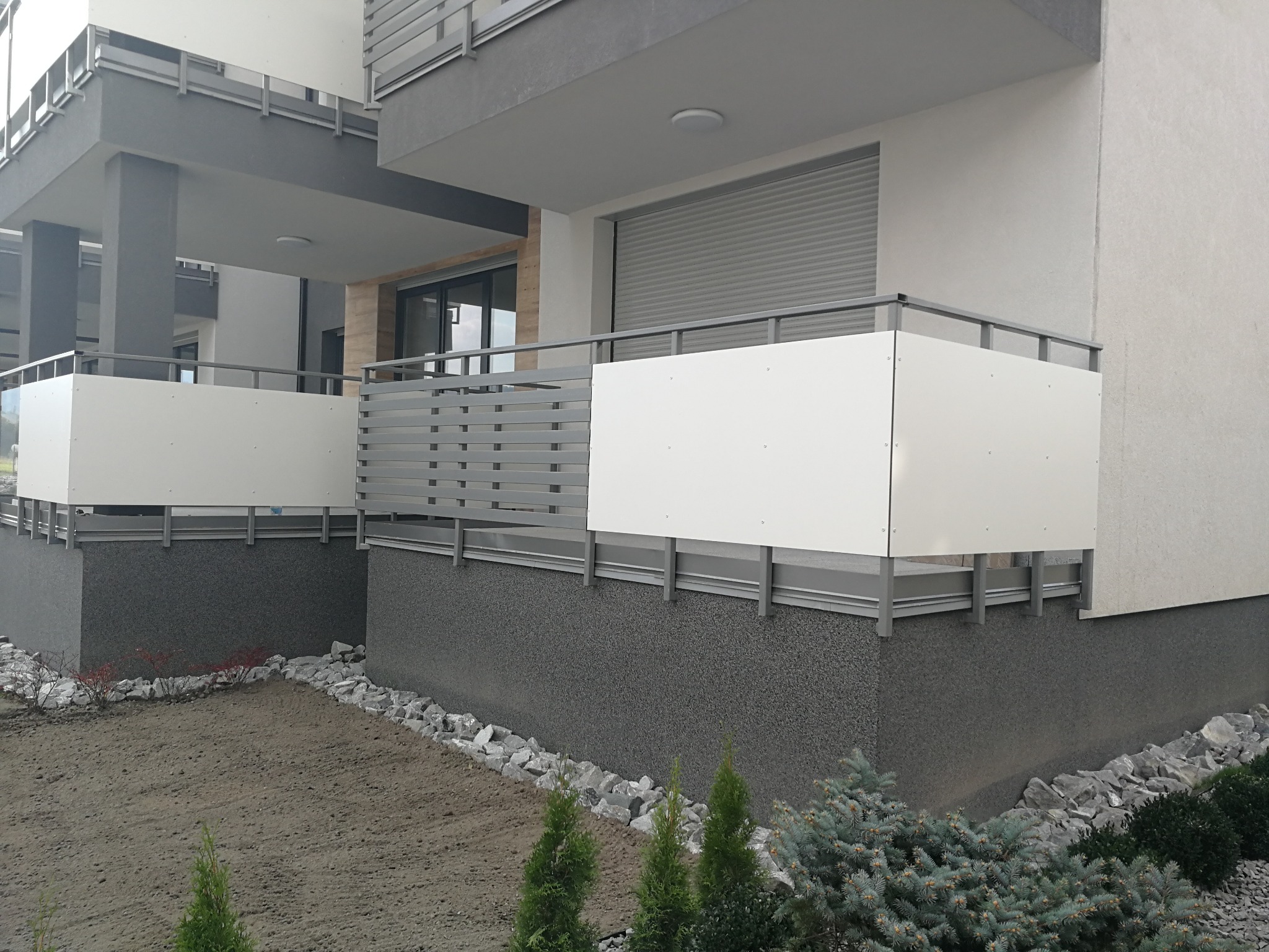 Nowoczesne balkony z szarymi, metalowymi barierkami i białymi panelami, widok z zewnątrz, z elementami zagospodarowania terenu.