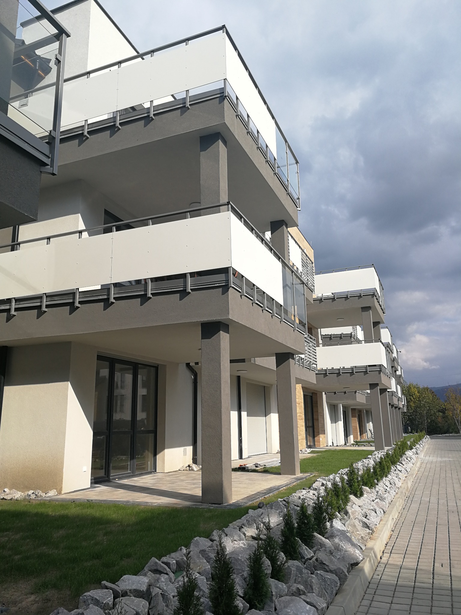 Nowoczesny budynek wielorodzinny z balkonami zabezpieczonymi panelami i szklanymi balustradami, widok z poziomu gruntu na elewację z elementami zieleni i kamieni.