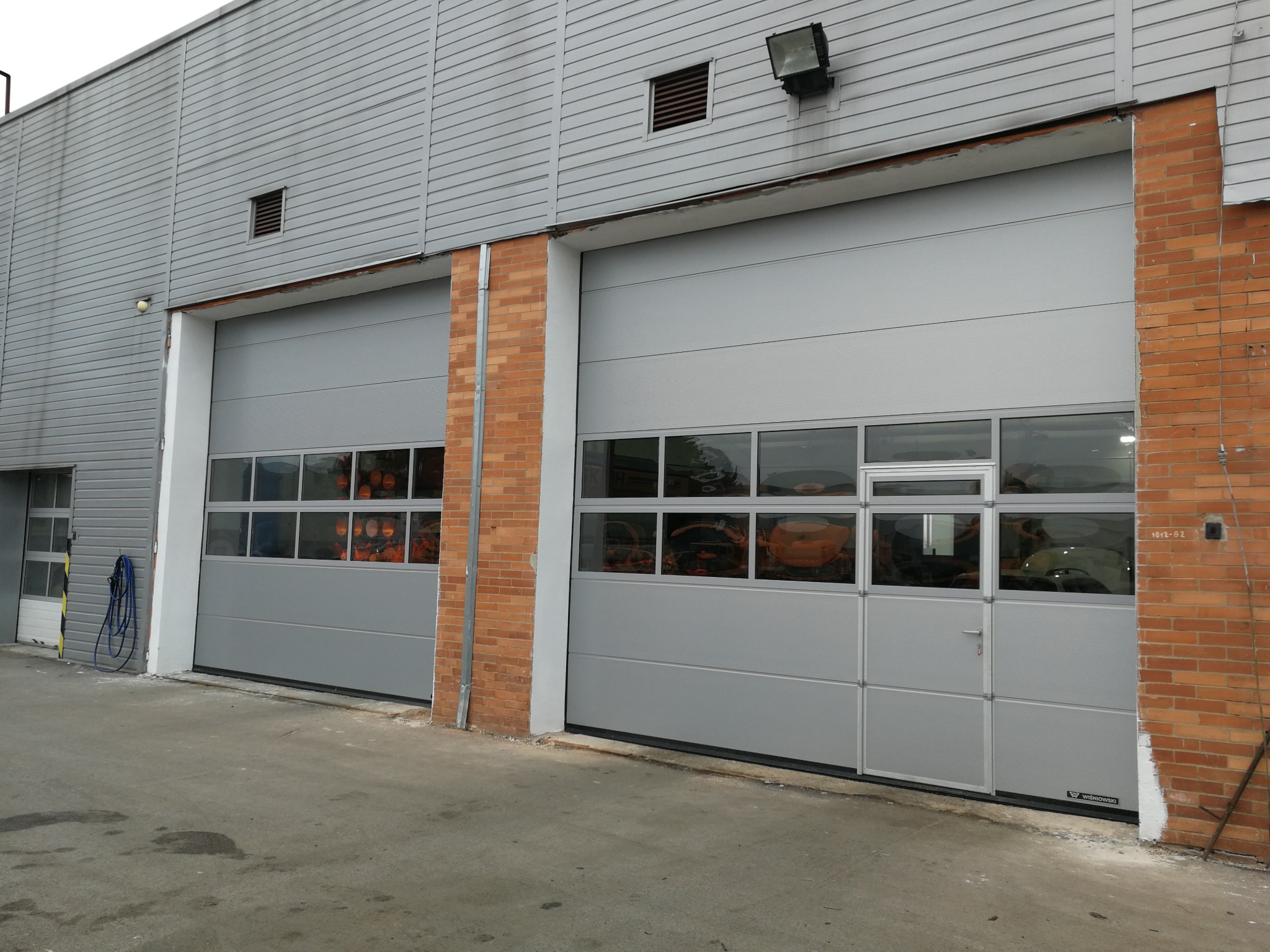 Dwie szare bramy segmentowe przemysłowe z oknami, umieszczone w budynku z szarą elewacją i ceglanymi elementami, widok z zewnątrz.