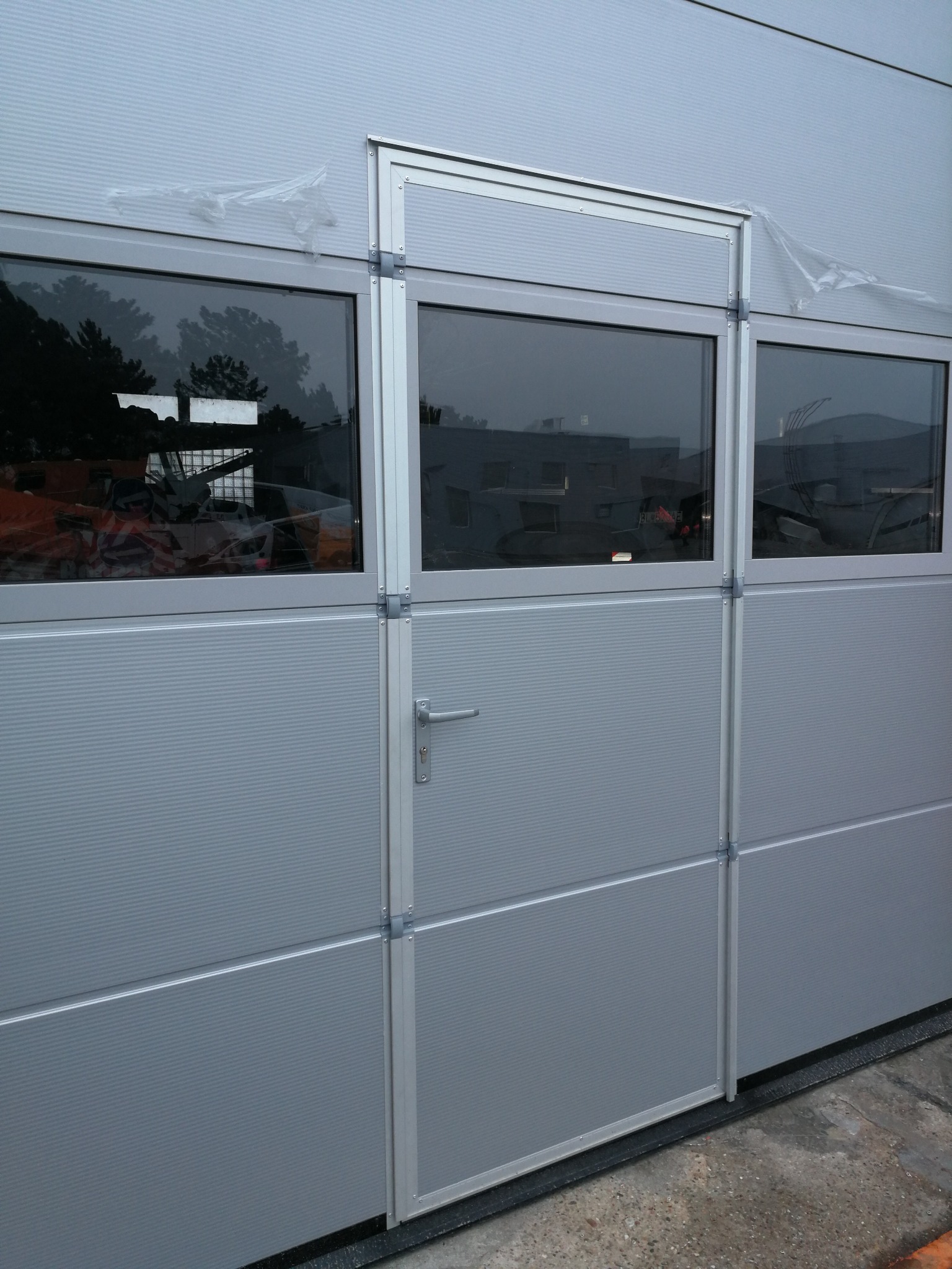 Szara brama przemysłowa segmentowa z oknami, widoczne elementy konstrukcyjne i uchwyt, z zewnątrz widoczne drzewa i zabudowania.