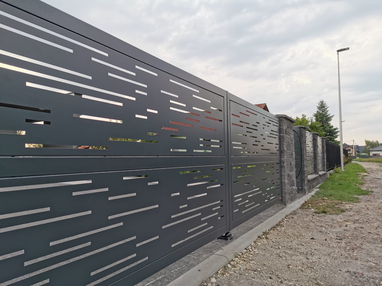 Nowoczesna, przesuwna brama wjazdowa z metalu w kolorze grafitowym, z poziomymi, prostokątnymi wycięciami, widoczna z boku, na tle posesji z kostką brukową i słupem oświetleniowym.