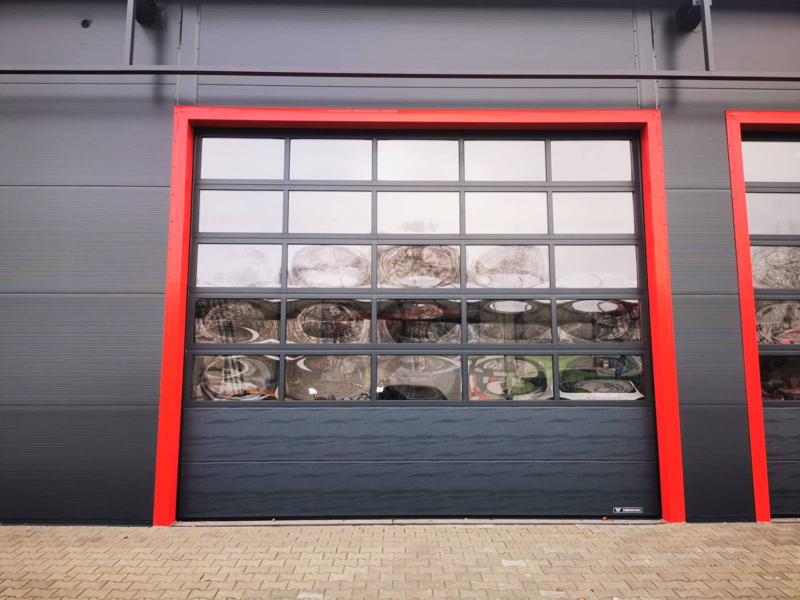 Szara segmentowa brama garażowa z czerwonym obramowaniem, podzielona na prostokątne okna, odbijająca otoczenie.