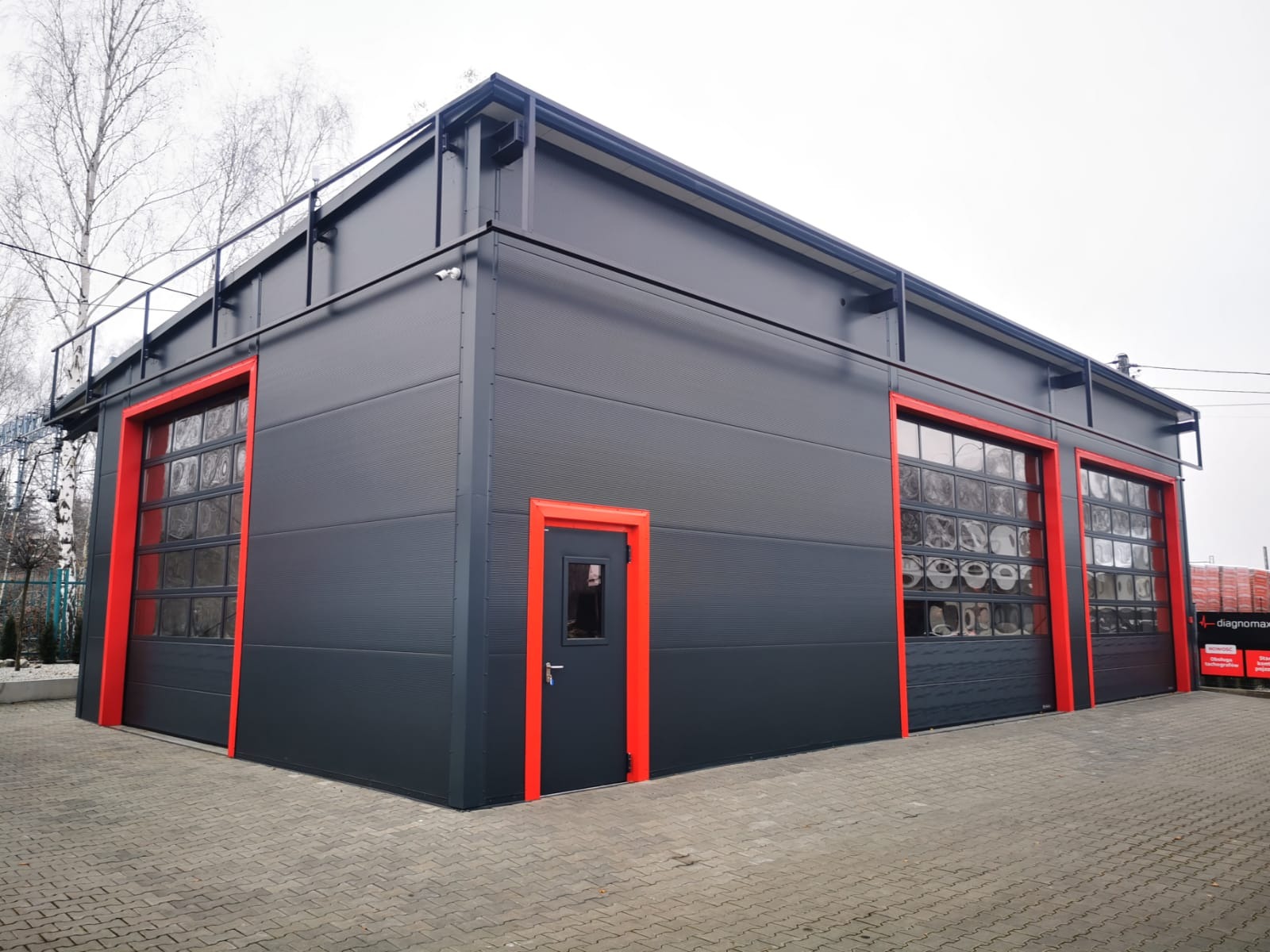 Szara hala przemysłowa z trzema bramami segmentowymi z oknami i drzwiami wejściowymi, obramowanymi na czerwono, widok z zewnątrz na kostce brukowej.