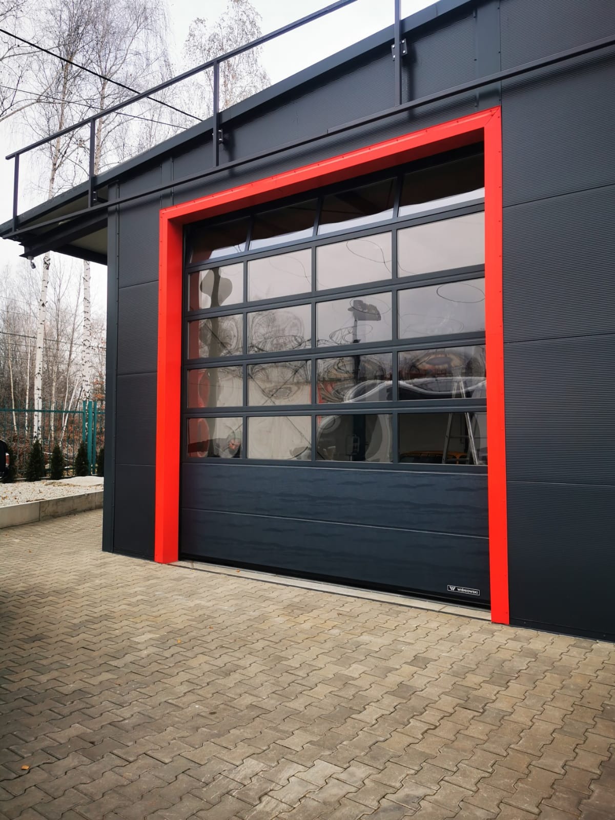 Szara brama garażowa segmentowa z przeszkleniami, obramowana czerwoną ramą, widoczna z zewnątrz budynku o szarej elewacji z poziomym wzorem, na pierwszym planie kostka brukowa.