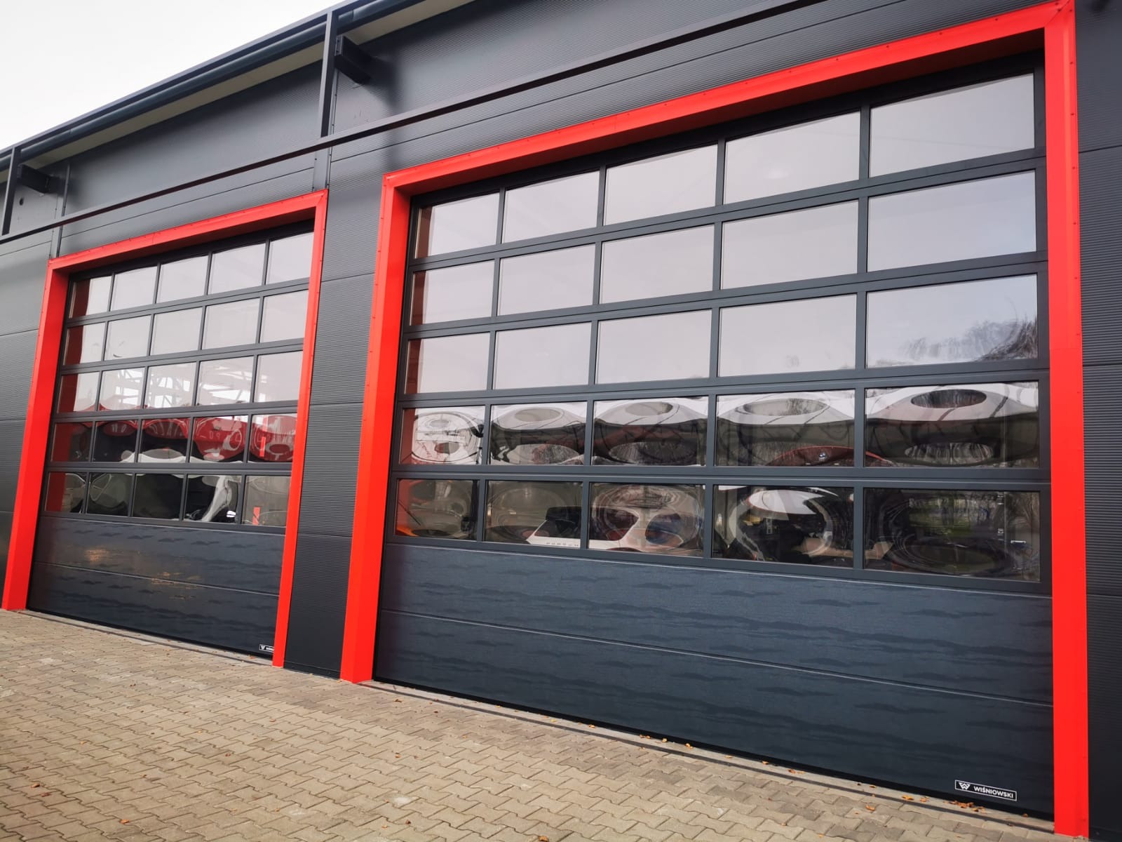 Dwie segmentowe bramy przemysłowe z przeszkleniami w czerwonych ramach, widoczne z zewnątrz budynku o szarej elewacji.