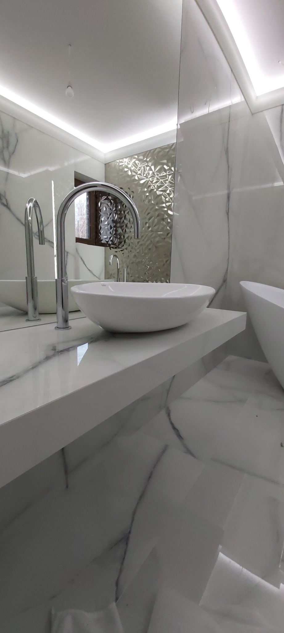 Nowoczesna łazienka z białą marmurową płytką, owalną umywalką nablatową i chromowaną baterią, oświetlenie LED podsufitowe.