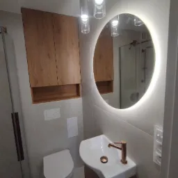 Nowoczesna łazienka z wiszącą szafką z drewnianymi frontami, okrągłym lustrem z podświetleniem LED, minimalistyczną umywalką z miedzianą baterią oraz wiszącą toaletą.