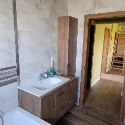 Nowoczesna łazienka z drewnopodobnymi płytkami na podłodze i ścianach, biała wanna i szafka pod umywalkę z drewnianym wykończeniem, widok na korytarz z drewnianymi schodami.