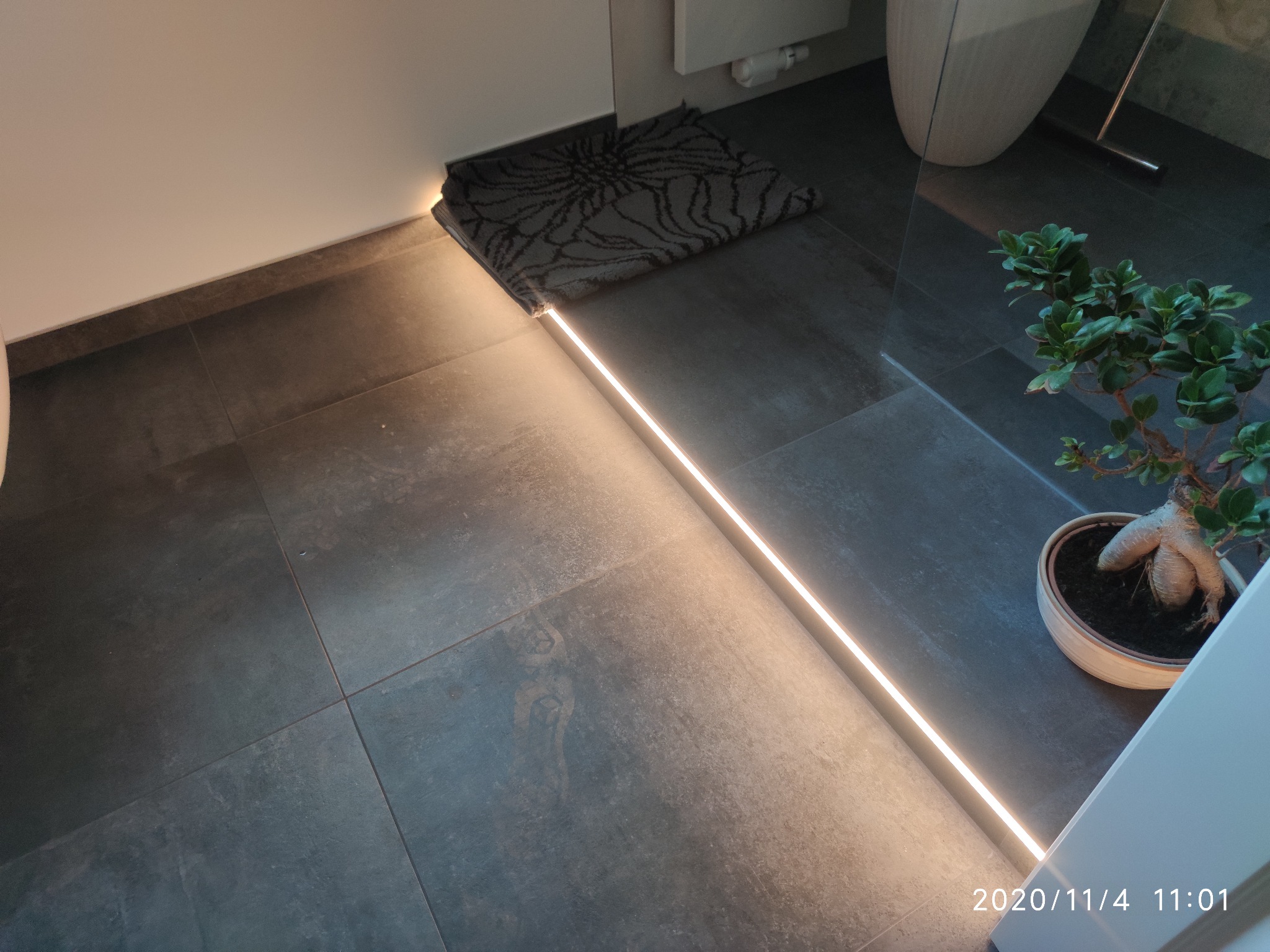 Nowoczesna łazienka z podświetlaną listwą przypodłogową LED, ciemnoszare płytki podłogowe, dywanik i bonsai w doniczce.