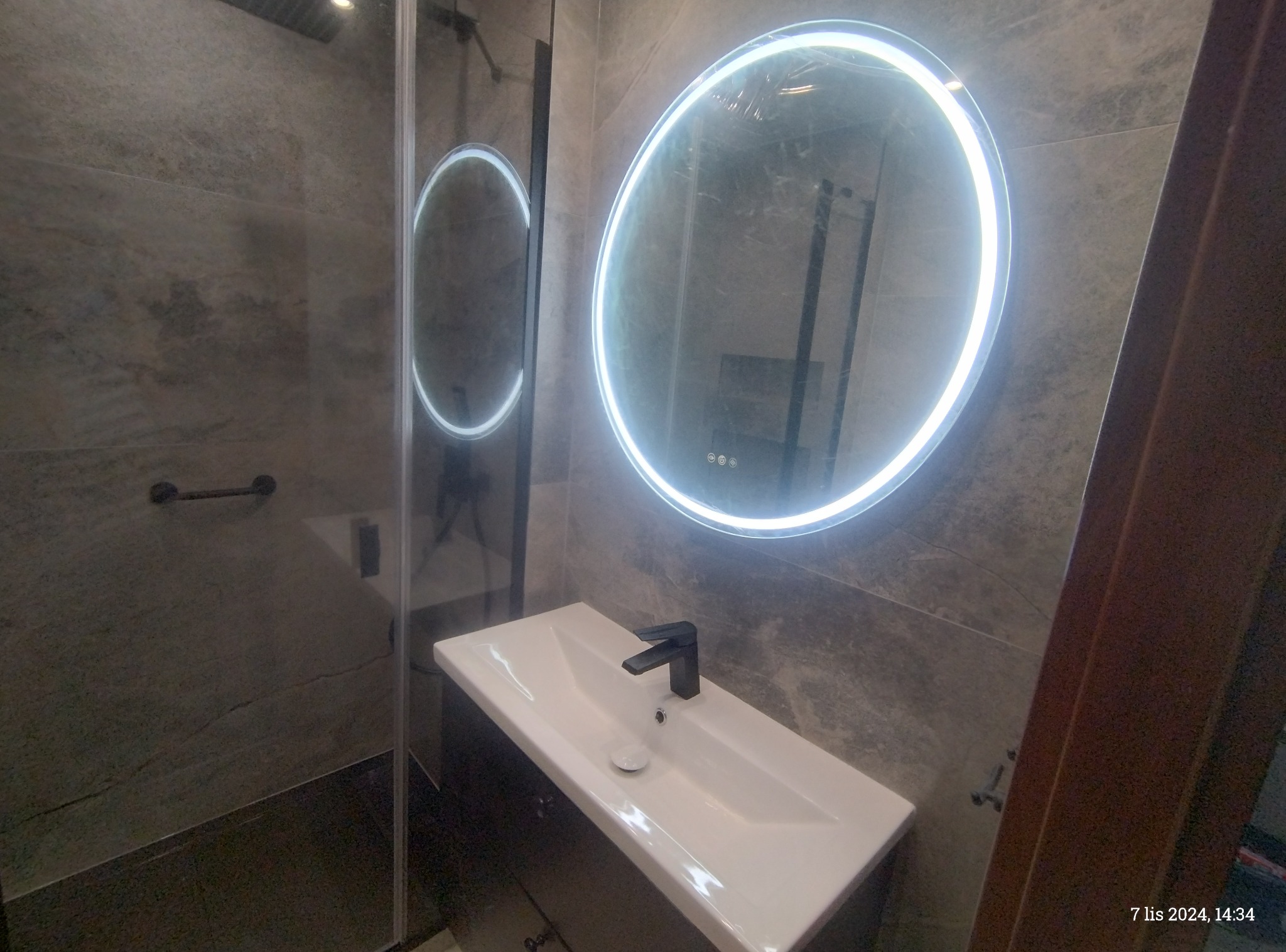 Nowoczesna łazienka z umywalką nablatową, czarną armaturą i okrągłym lustrem z podświetleniem LED na tle szarych płytek imitujących kamień.