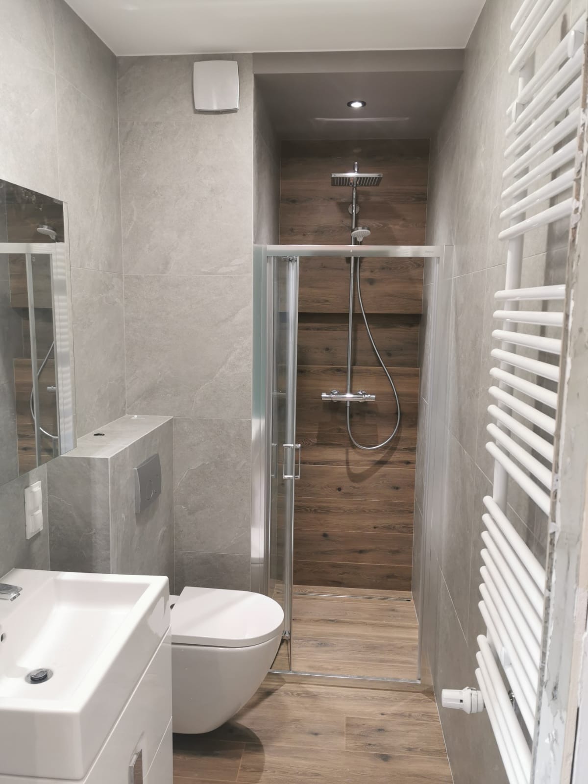Nowoczesna łazienka z kabiną prysznicową wyłożoną drewnopodobnymi płytkami, szara glazura na ścianach, biała umywalka i toaleta, grzejnik drabinkowy.
