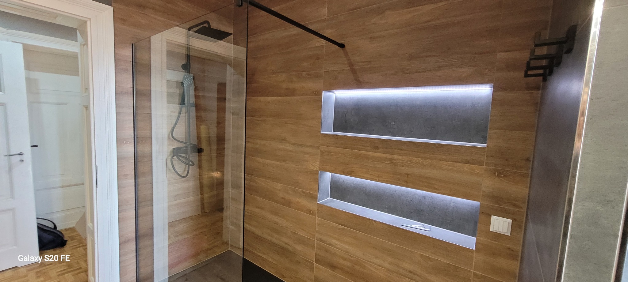 Nowoczesna łazienka z kabiną prysznicową typu walk-in, wykończona płytkami imitującymi drewno, z wnękami podświetlonymi LED i czarną armaturą.