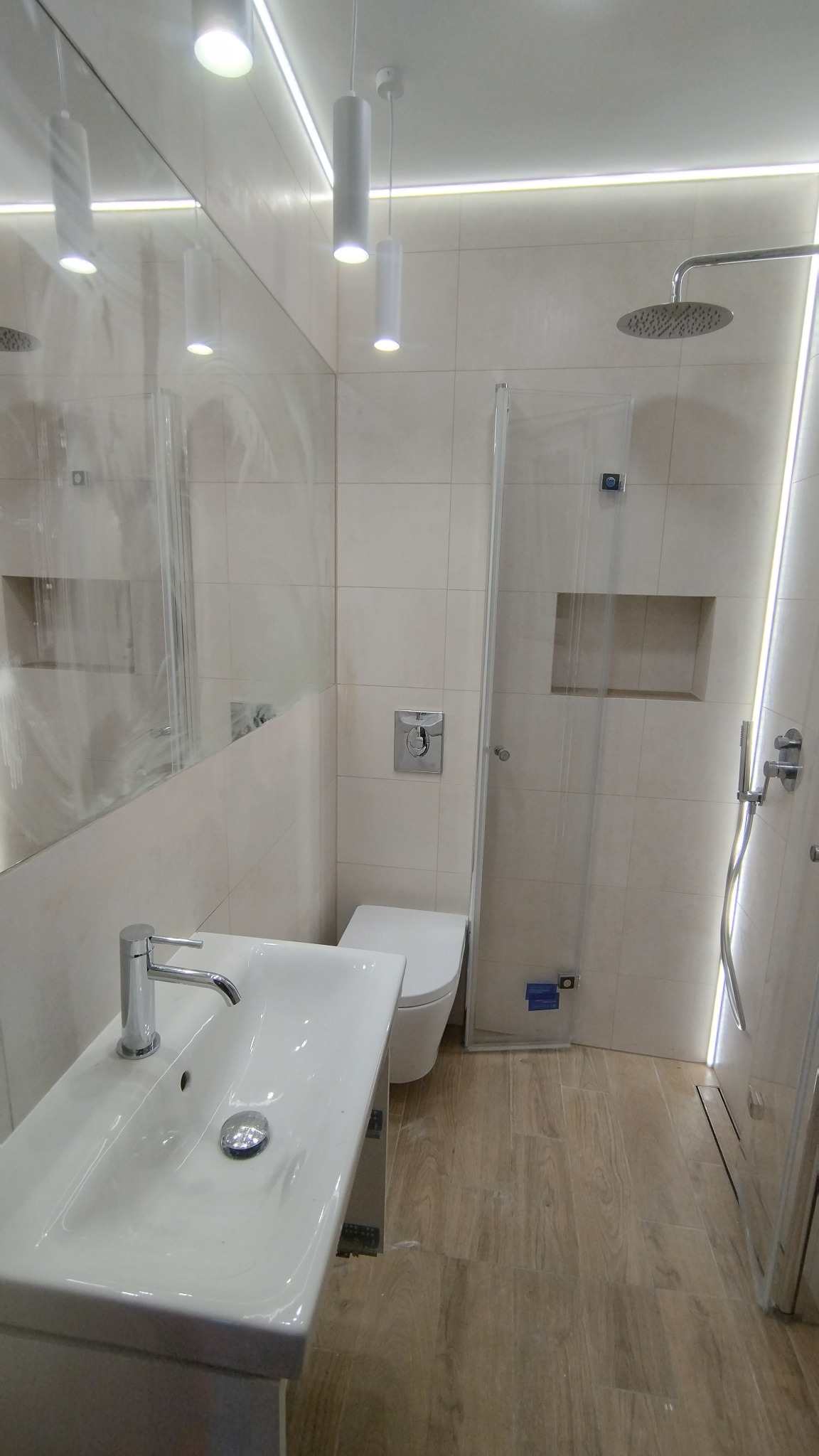 Nowoczesna łazienka z białymi płytkami, kabiną prysznicową typu walk-in, podwieszaną toaletą i umywalką nablatową z chromowaną armaturą, oświetlona listwami LED i cylindrycznymi lampami wiszącymi.