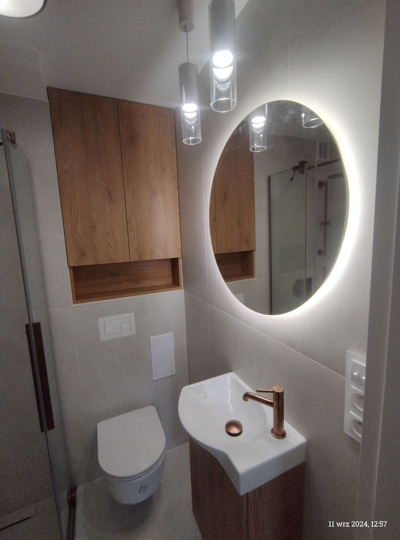 Nowoczesna łazienka z wiszącą szafką z drewnianymi frontami, okrągłym lustrem z podświetleniem LED, minimalistyczną umywalką z miedzianą baterią oraz wiszącą toaletą.