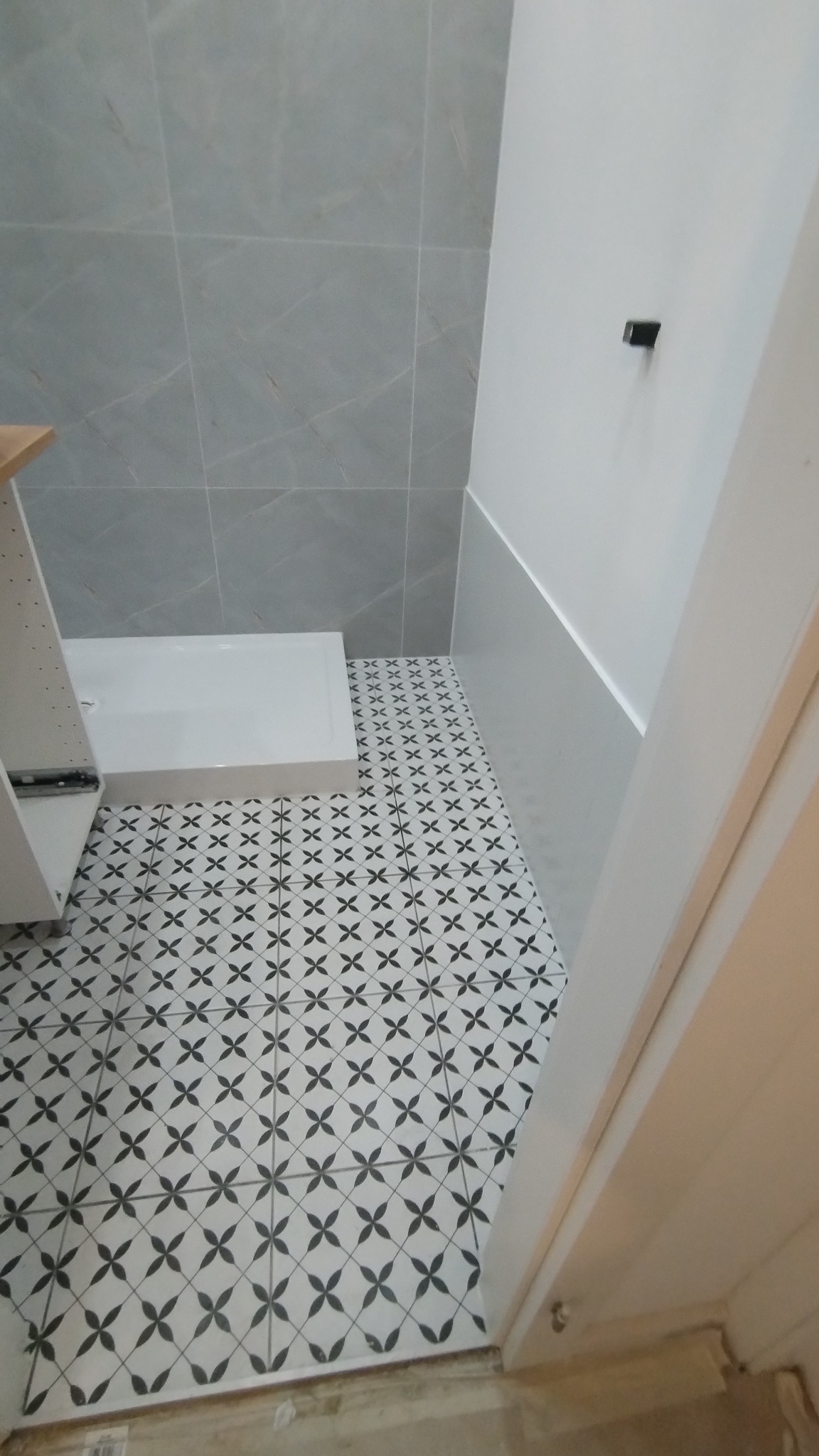 Fragment łazienki z ułożonymi płytkami podłogowymi w czarno-biały wzór kwiatowy, widoczny brodzik i szare płytki ścienne.