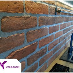 Płytkarz Wrocławski - Ściana wykończona cegłą klinkierową w kuchni, z widocznym fragmentem zlewu i baterii oraz relingiem na ręczniki. Logo firmy glazurniczej w lewym dolnym rogu.