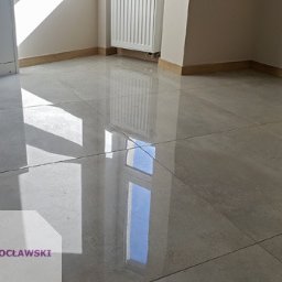 Płytkarz Wrocławski - Błyszcząca, szara podłoga z płytek w pustym pokoju, odbijająca światło okna i lampy, widoczne listwy przypodłogowe i grzejnik.