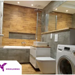 Nowoczesna łazienka z drewnopodobnymi płytkami na ścianie, białą armaturą i pralką, widoczny wnękowy schowek nad wanną.