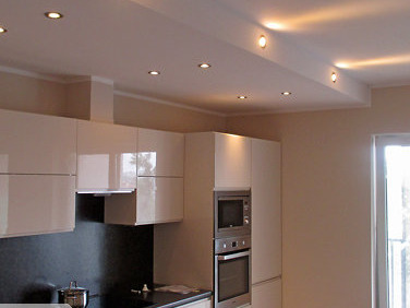 Jasna kuchnia z białymi, lakierowanymi szafkami, czarnym blatem i podwieszanym sufitem z punktowym oświetleniem LED.