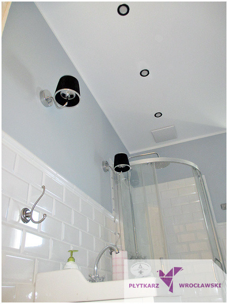 Jasna łazienka z białymi, prostokątnymi płytkami na ścianach, kabiną prysznicową z zaokrąglonymi drzwiami i umywalką z baterią retro oraz czarnymi lampami ściennymi.