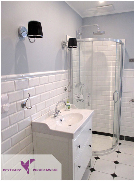 Biała umywalka z szafką, kabina prysznicowa z chromowaną armaturą i czarne, abażurowe kinkiety w wykończonej łazience z białymi, cegiełkowymi płytkami ściennymi i wzorzystą podłogą.