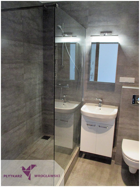 Nowoczesna łazienka z szarymi płytkami imitującymi kamień, kabiną prysznicową typu walk-in z odpływem liniowym, białą szafką pod umywalką i lustrem z oświetleniem LED.