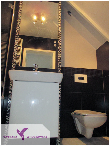 Nowoczesna łazienka na poddaszu z czarnymi płytkami, lustrem obramowanym mozaiką i podwieszaną toaletą.