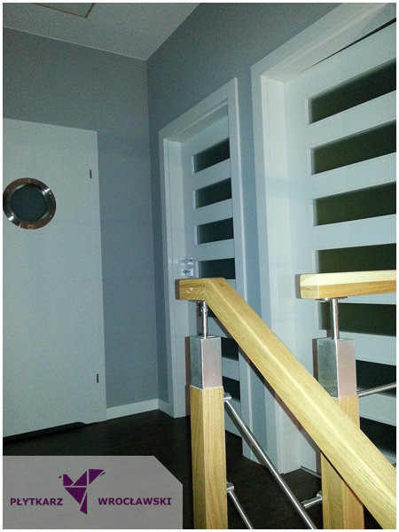 Szare wnętrze z dwiema białymi drzwiami: jedne z okrągłym oknem w stylu bulaja, drugie z poziomymi przeszkleniami, widoczna drewniana balustrada schodów z metalowymi elementami.