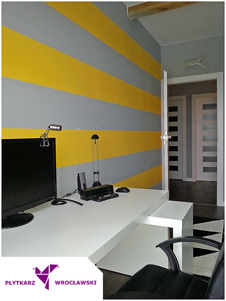 Pokój z biurkiem i ścianą w poziome, szaro-żółte pasy. W tle widoczne otwarte drzwi i dekoracja w kształcie samolotu.
