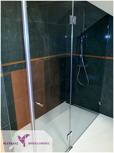 Nowoczesna kabina prysznicowa z transparentnymi drzwiami i czarnymi płytkami, akcentowana poziomym paskiem w kolorze pomarańczowym, widoczny deszczownica i bateria prysznicowa.