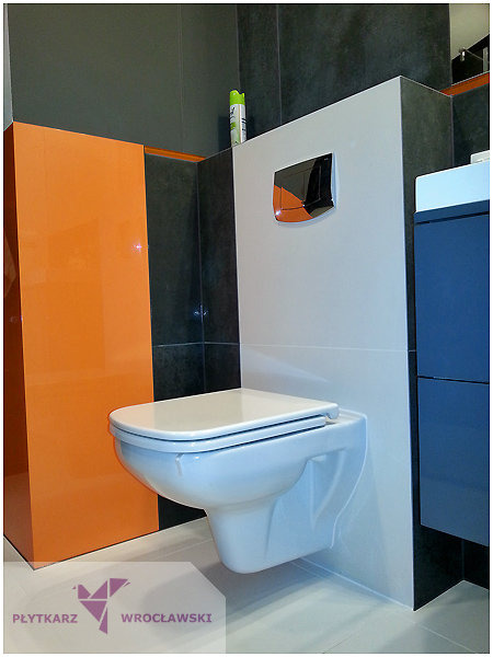 Nowoczesna, podwieszana toaleta z chromowanym przyciskiem spłukującym, obok szafki w kolorze pomarańczowym i granatowym na tle szarych płytek.