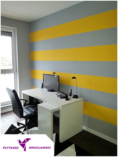 Pokój biurowy z pomalowaną ścianą w poziome pasy szaro-żółte, białe biurko z komputerem i czarne krzesło obrotowe.