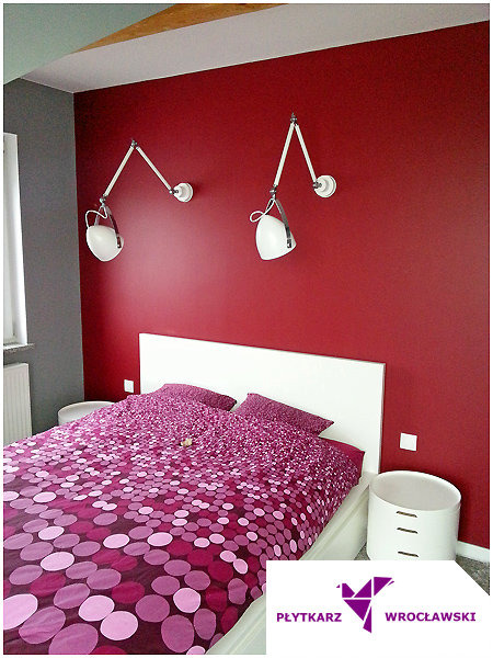 Pokój z czerwonymi ścianami, białą ramą łóżka i szafkami nocnymi, oświetlony dwoma białymi lampami ściennymi z regulowanymi ramionami.