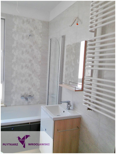 Nowoczesna łazienka z geometrycznymi płytkami, kabiną prysznicową za umywalką, grzejnikiem drabinkowym i lustrem z półką. Widoczny fragment wanny z baterią.