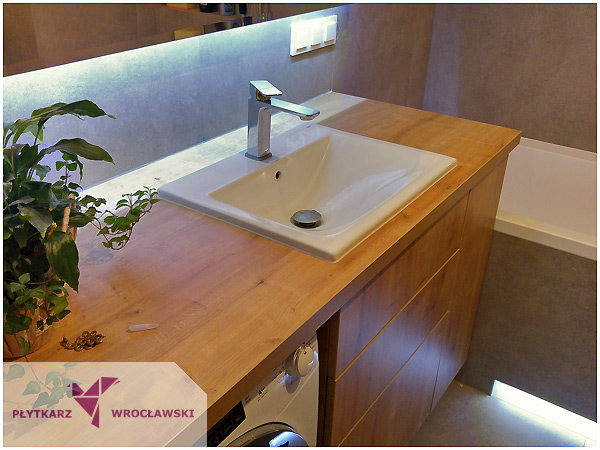 Nowoczesna łazienka: drewniany blat z umywalką nablatową i baterią, podświetlane lustro, pralka w zabudowie, roślina doniczkowa, subtelne oświetlenie pod wanną.