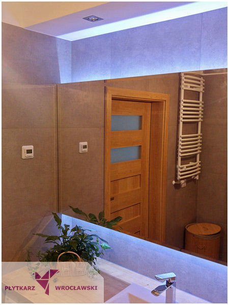 Nowoczesna łazienka z szarymi płytkami, drewnianą ramą drzwi i podświetlanym lustrem, odbijającym grzejnik drabinkowy i roślinę w koszyku.