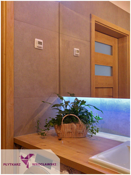 Wykończona łazienka z szarymi płytkami, drewnianą ramą drzwi i blatem z koszykiem z roślinami, podświetlona listwą LED i dwoma termostatami na ścianie.