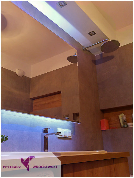 Nowoczesna łazienka z szarymi płytkami, podwójnym prysznicem deszczownicą i podświetleniem LED, widoczny fragment blatu z logo firmy.