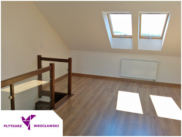 Poddasze z drewnianą podłogą, dwoma oknami dachowymi, białym grzejnikiem i balustradą ze szkła i drewna.