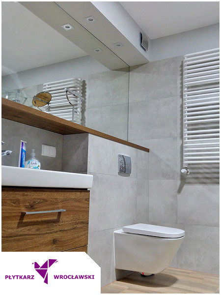 Nowoczesna łazienka z szarymi płytkami wielkoformatowymi, podwieszaną miską WC, drewnianą szafką pod umywalką i grzejnikiem drabinkowym.