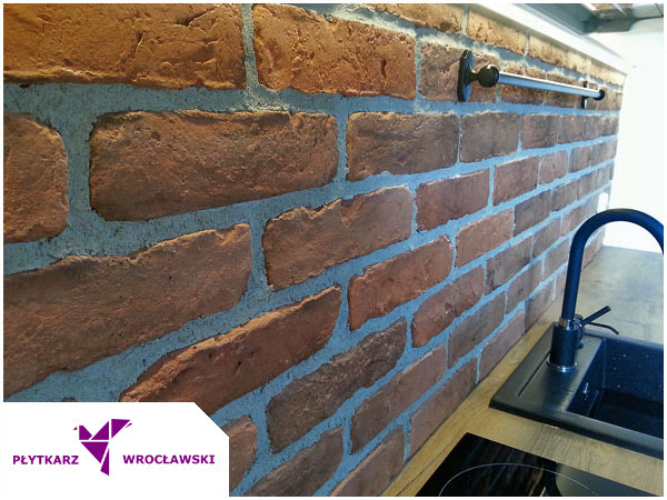 Ściana z cegły klinkierowej z szarą fugą w kuchni we Wrocławiu, widoczny fragment blatu, zlew i bateria.