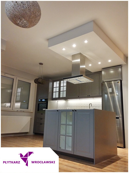 Szara kuchnia z wyspą, stalowym okapem i oświetleniem LED podszafkowym, widok na wykończone wnętrze z drewnianą podłogą.