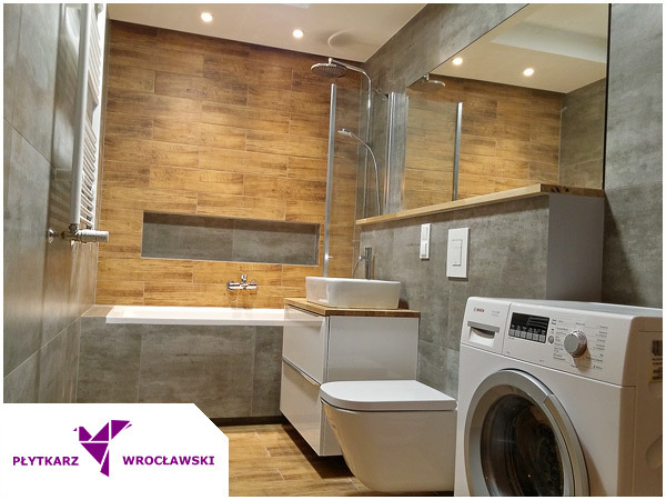 Nowoczesna łazienka z drewnopodobnymi płytkami na ścianie, białą armaturą i pralką, widoczny wnękowy schowek nad wanną.