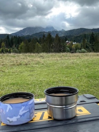 Dwa metalowe kubki termiczne z kawą na czarnej skrzynce narzędziowej DeWalt, w tle zielona łąka, las i góry pod pochmurnym niebem.