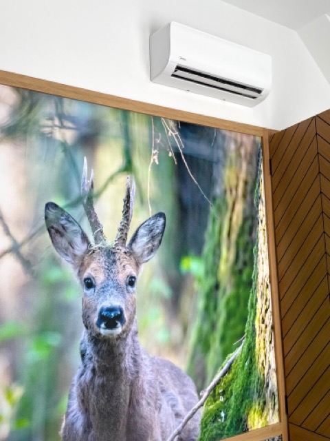 Biały klimatyzator Midea zamontowany nad obrazem jelenia w drewnianej ramie, z drewnianą ścianą w tle.
