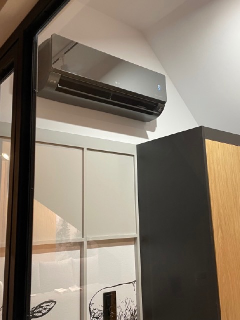 Szary klimatyzator marki LG zamontowany na białej ścianie w rogu pokoju, widoczny fragment szafki z drewnianymi drzwiczkami i dekoracyjny panel ścienny z czarnym wzorem.