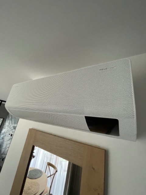 Biała jednostka wewnętrzna klimatyzatora typu split, model WindFree, zamontowana na ścianie nad lustrem w drewnianej ramie, odbijającym fragment jadalni.