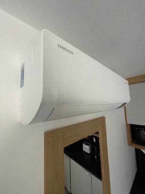 Biała jednostka klimatyzacji Samsung zamontowana na ścianie w mieszkaniu, widok od dołu, z fragmentem kuchni w tle.