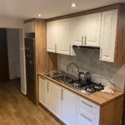 Nowoczesna kuchnia z białymi frontami szafek i drewnianym blatem, srebrne uchwyty, płyta gazowa z garnkiem, marmurowa ściana nad blatem, drewniana podłoga.